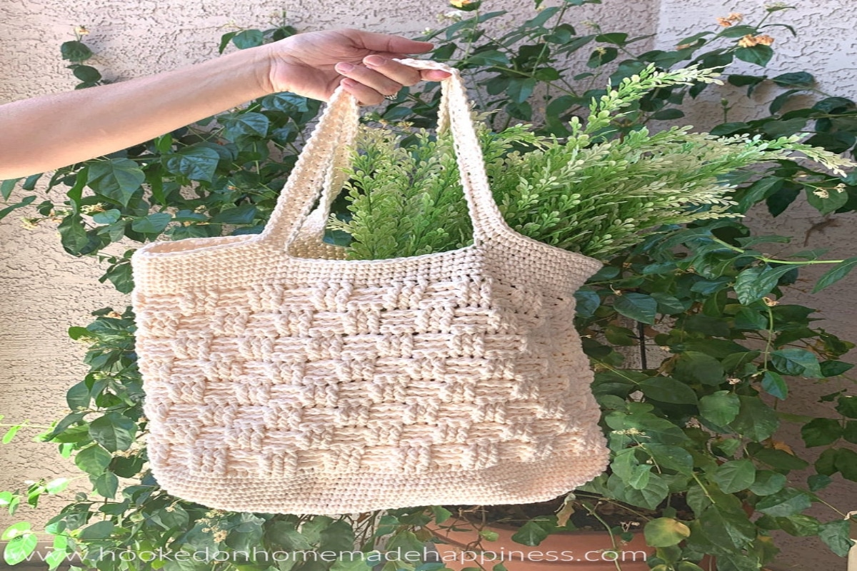 A cream basketweave crochet bag with thick handles and leaves poking out in front of a large house plant.