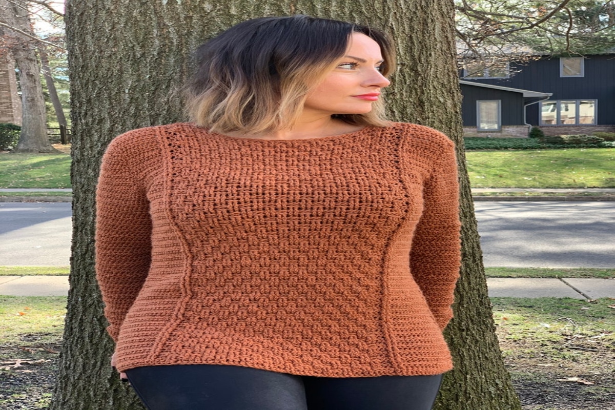 A woman standing in front of a tree wearing a light orange crochet sweater with a basketweave panel down the middle.