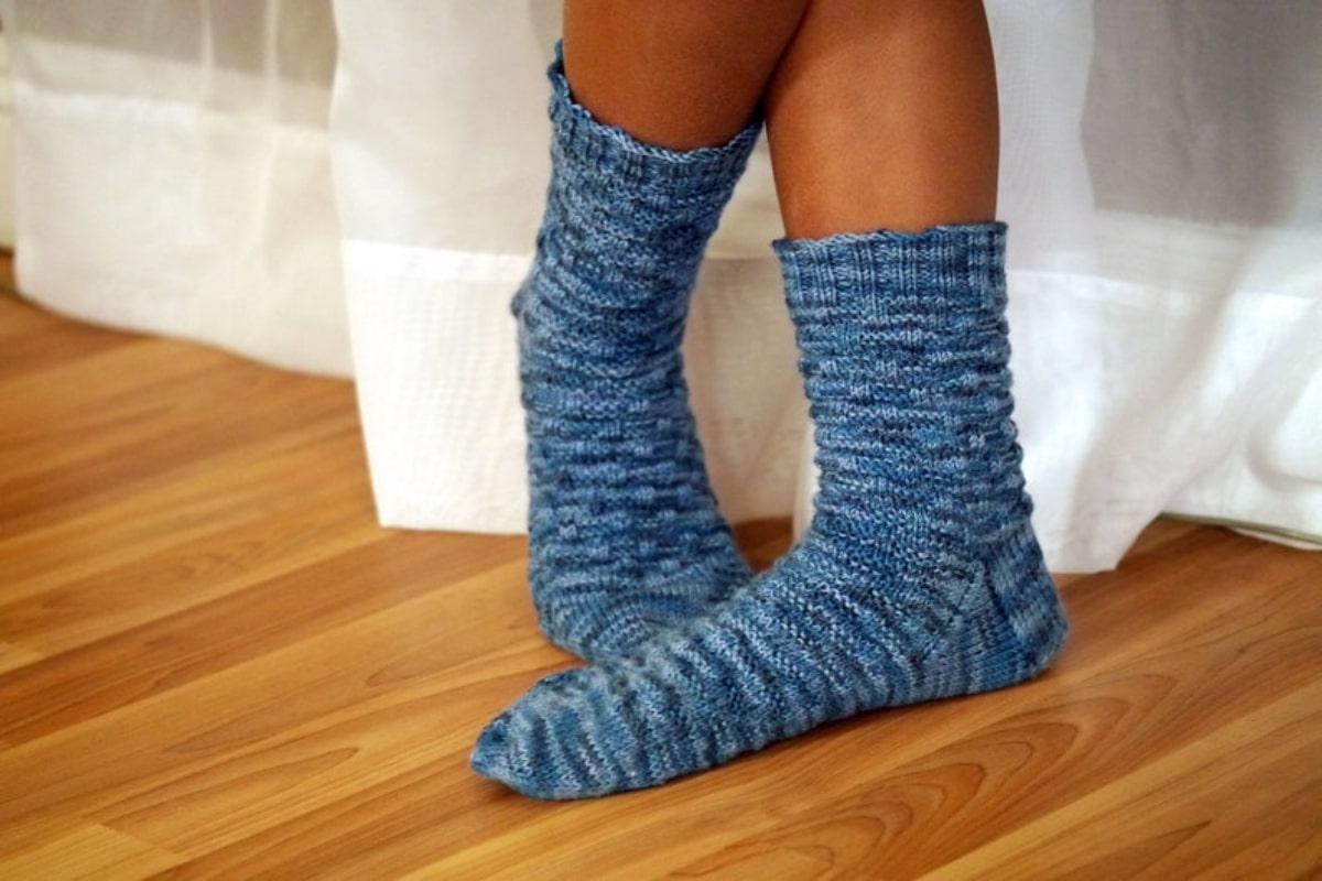 A woman’s feet in a pair of blue basketweave crochet socks standing on a wooden floor by some white curtains.