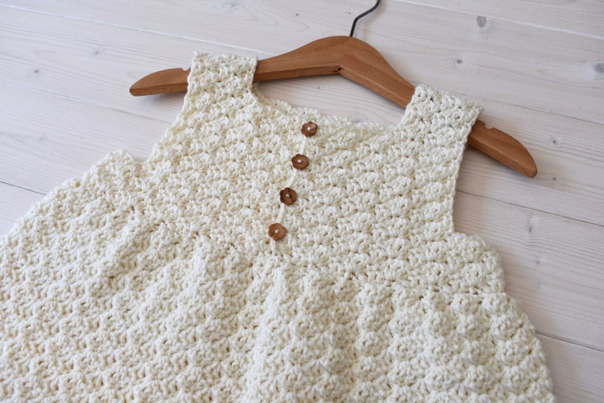 White crochet sleeveless baby dress with thick straps and four small brown buttons down the middle on a wooden hanger.