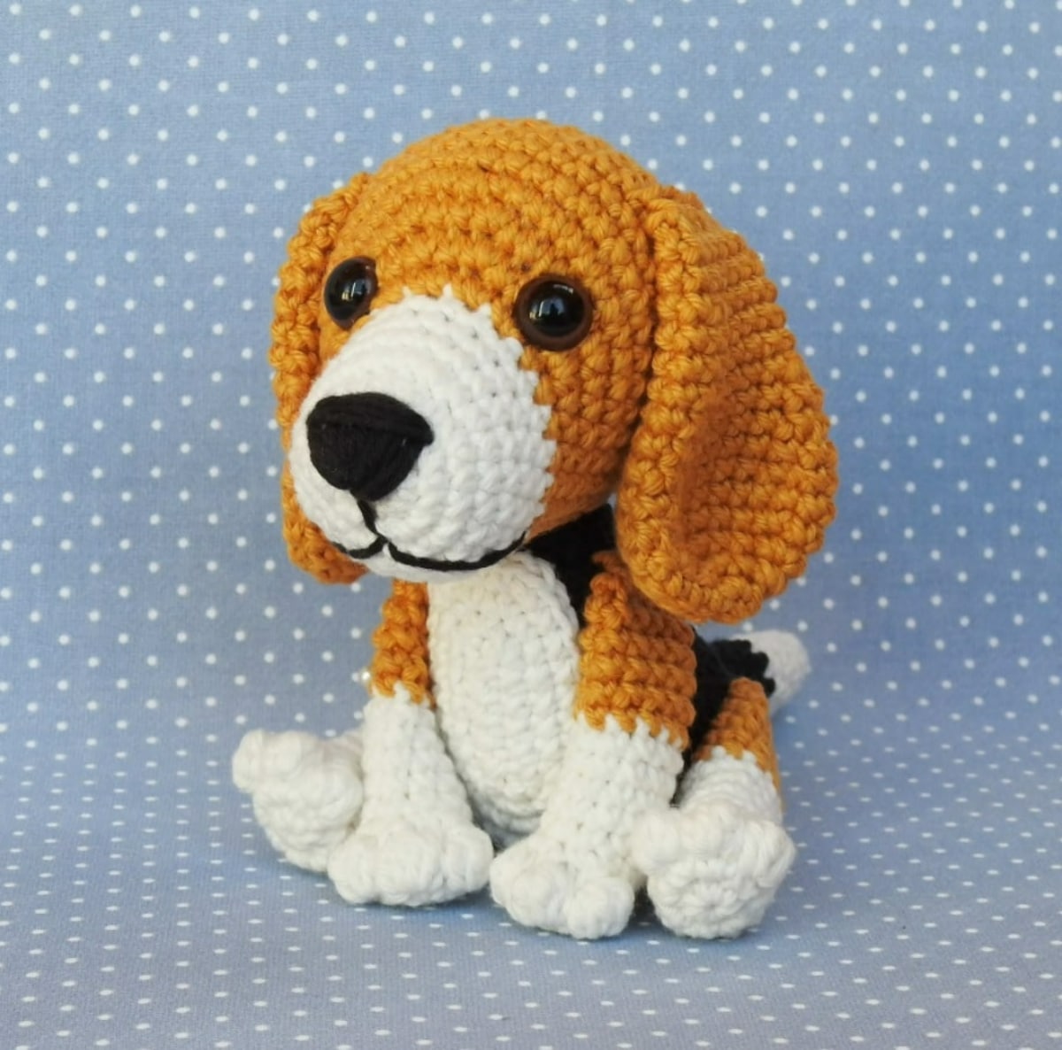 A crochet stuffed Beagle with a brown face and ears and white body, nose, and legs sitting on a blue and white spotty background.