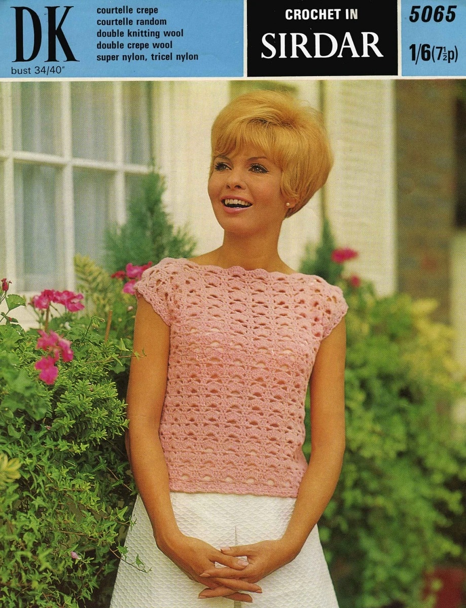 Blond woman wearing a 1960s style pink crochet t-shirt with white bottoms in front of a pink flowery shrub.