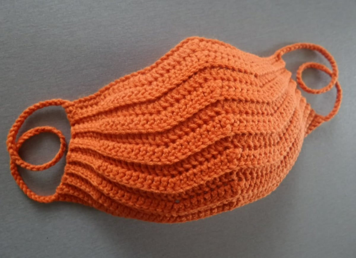 Orange crochet face mask with thin orange straps to place around the ears on a gray background.
