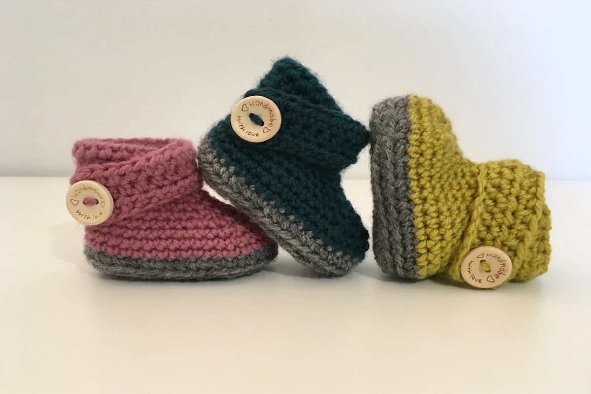Pink crochet baby boot with a strap and button to secure next to a black and yellow boot with the same style and gray soles.