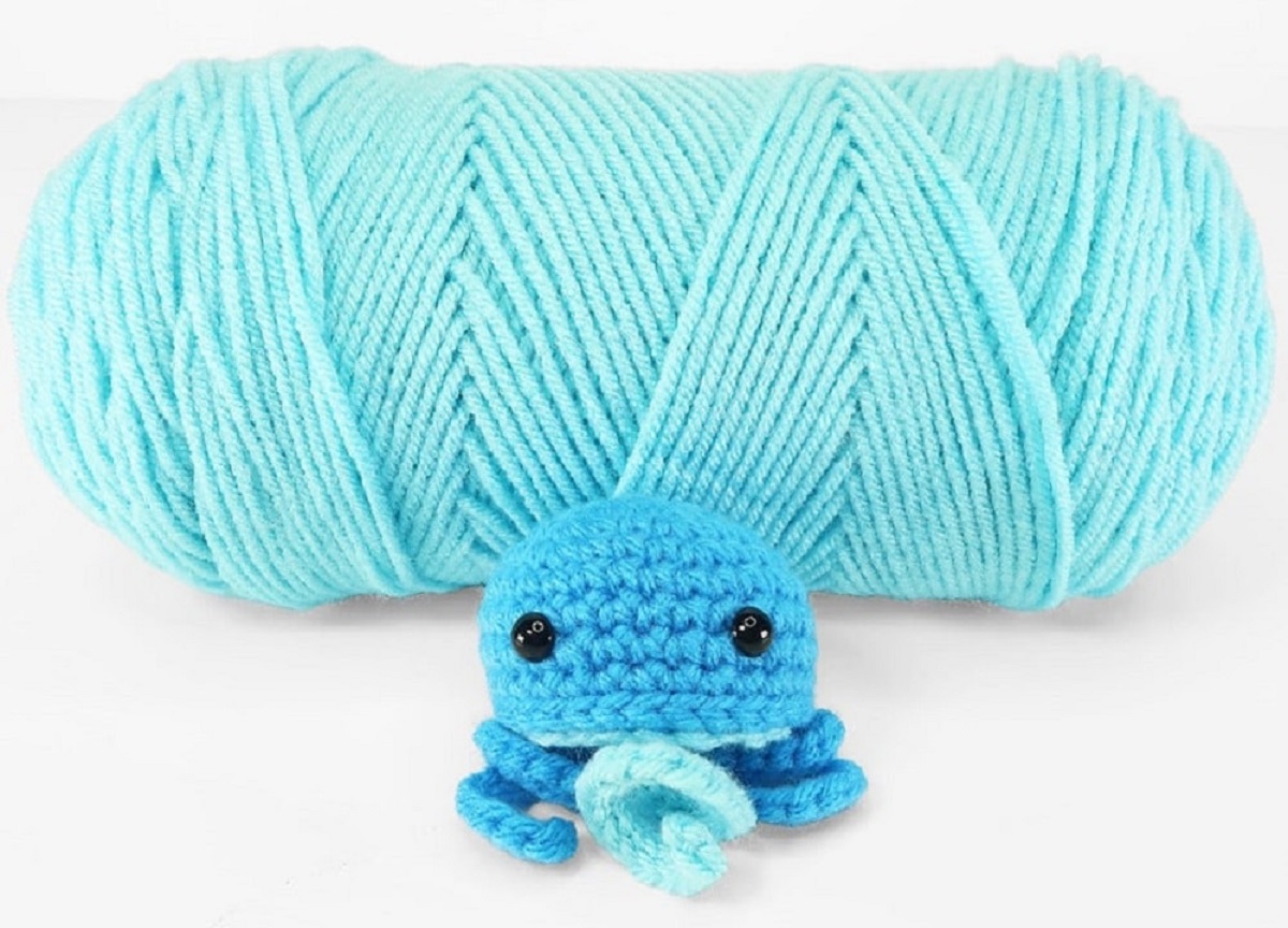 Miniature blue crochet jellyfish with one light blue tentacle in the center sitting in front of a blue ball of yarn.