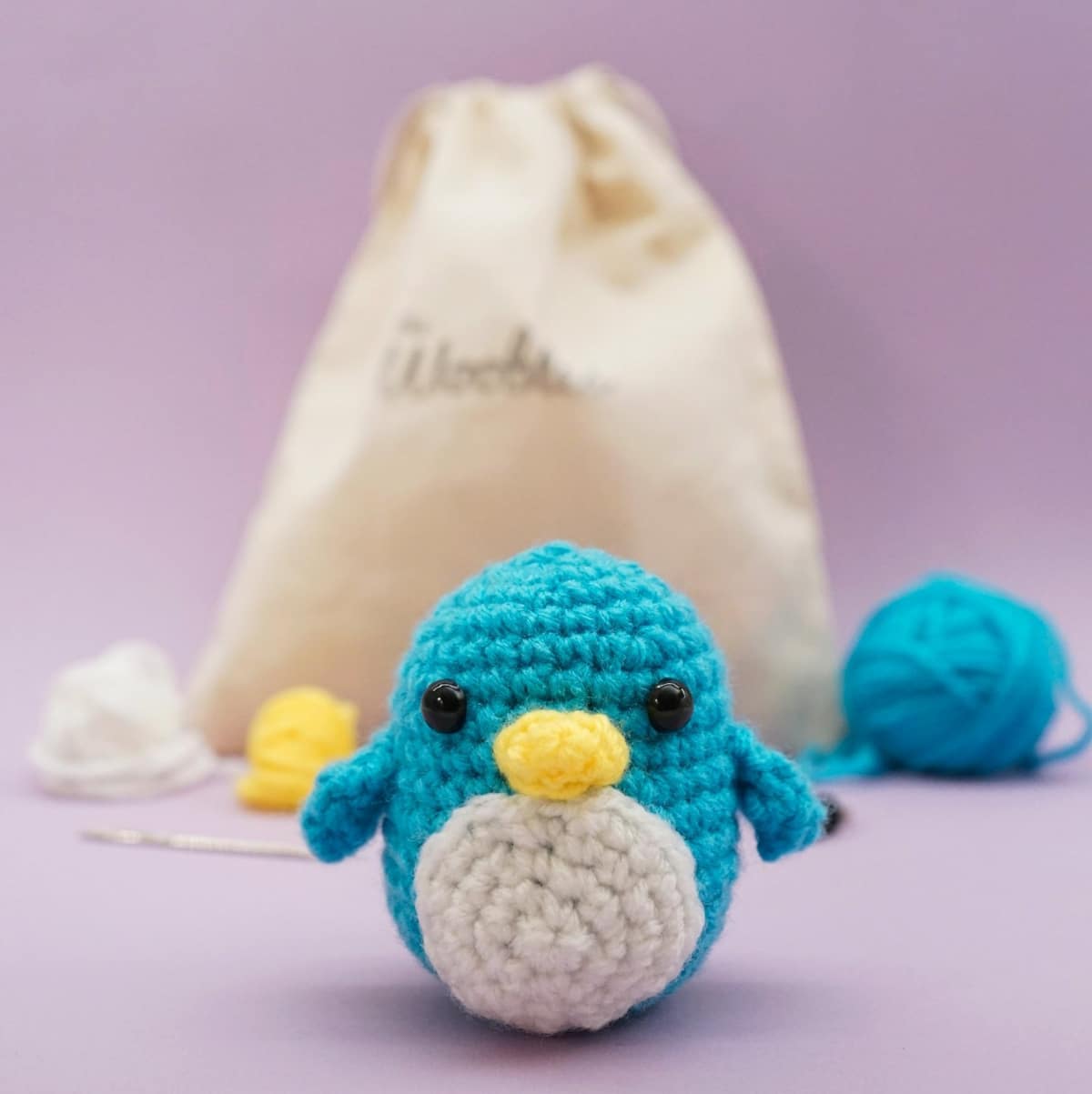 Small crochet blue penguin with a white chest, yellow nose, and black eyes in front of a pink background, brown bag, and balls of yarn.