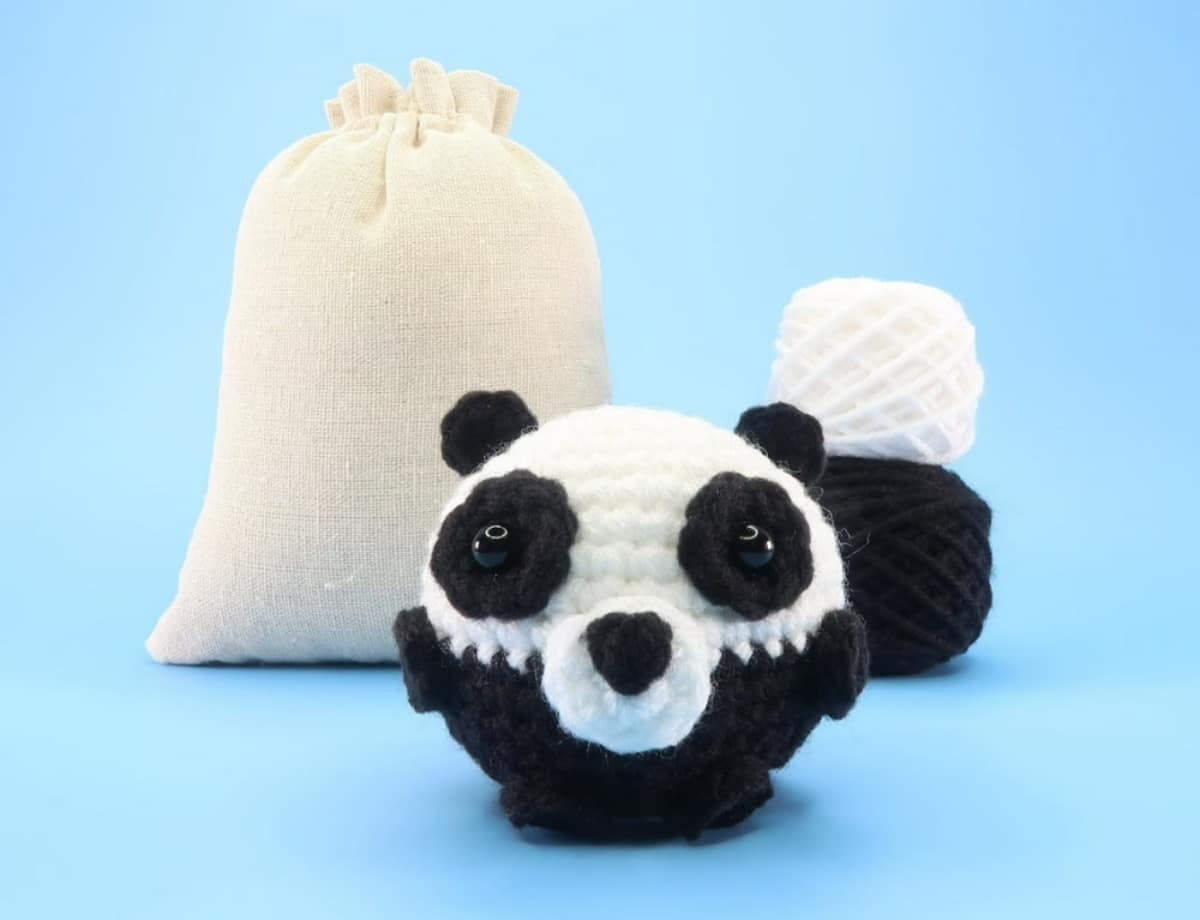 Small ball shaped black and white panda face with black and white yarn balls stacked behind it and a cream bag next to them.