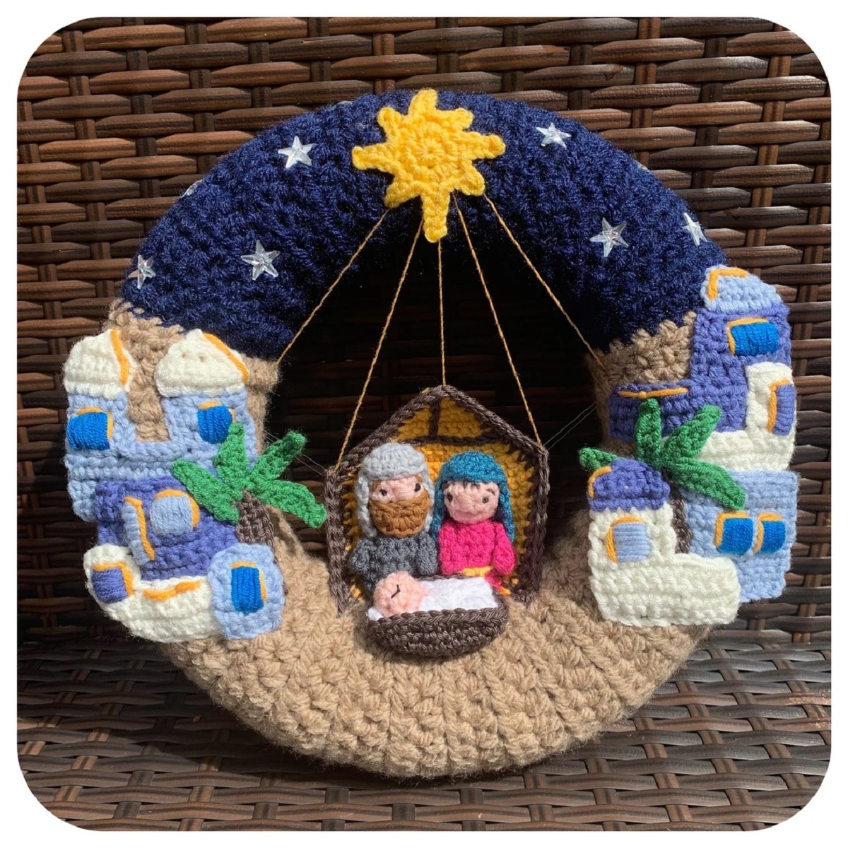 A crochet wreath with a blue night sky and yellow star in the center, Bethlehem on the side, and Mary, Joseph and Jesus in the center.