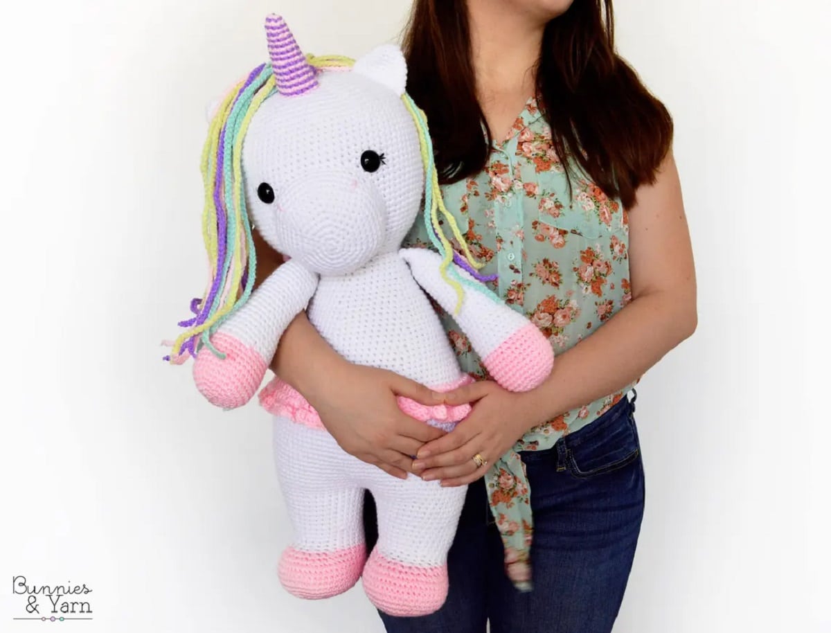 A woman holding a large white crochet unicorn with pink feet, a purple horn, and a purple and green mane.