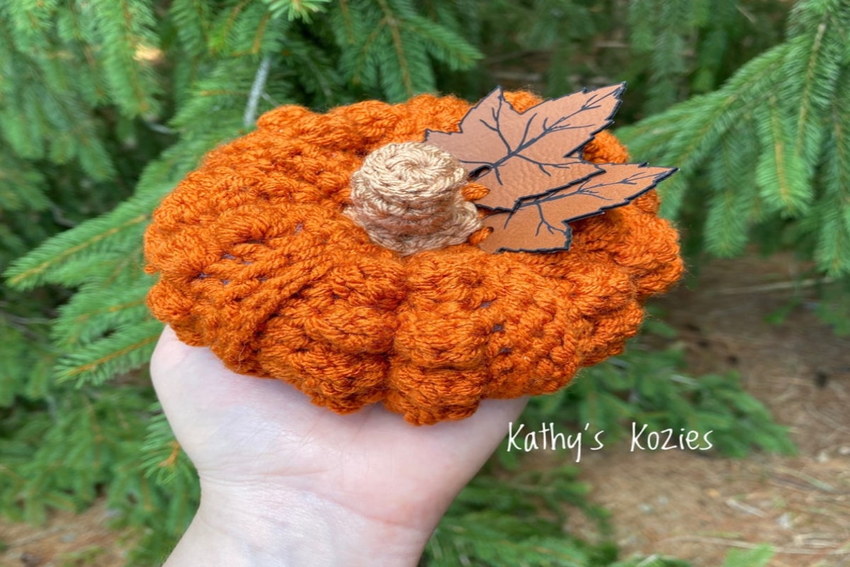 A hand holding a small orange crochet pumpkin with a brown stem in the middle and two brown leaves on the side.