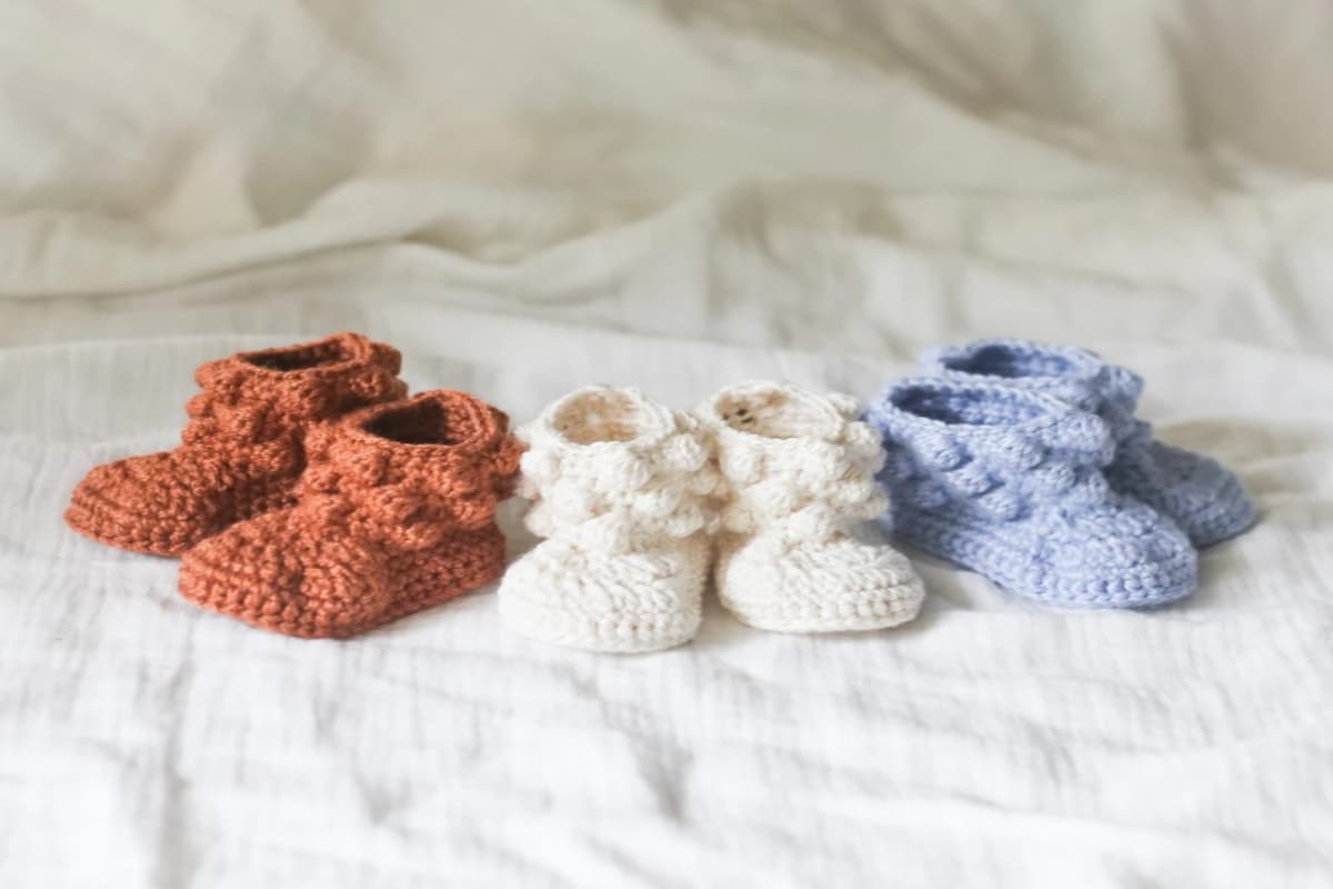 Three pairs of crochet baby booties with bobbles around the ankles. Boots are rust colored, cream, and baby blue on a white blanket.