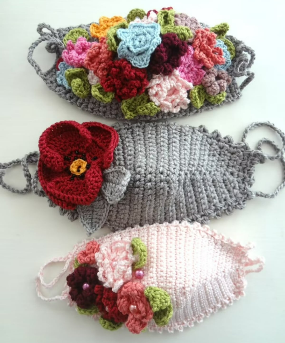 A pale pink crochet face mask with 3-D flowers on the side, a gray face mask with a large flower on the side, and a gray facemask with 3-D flowers all over the front.