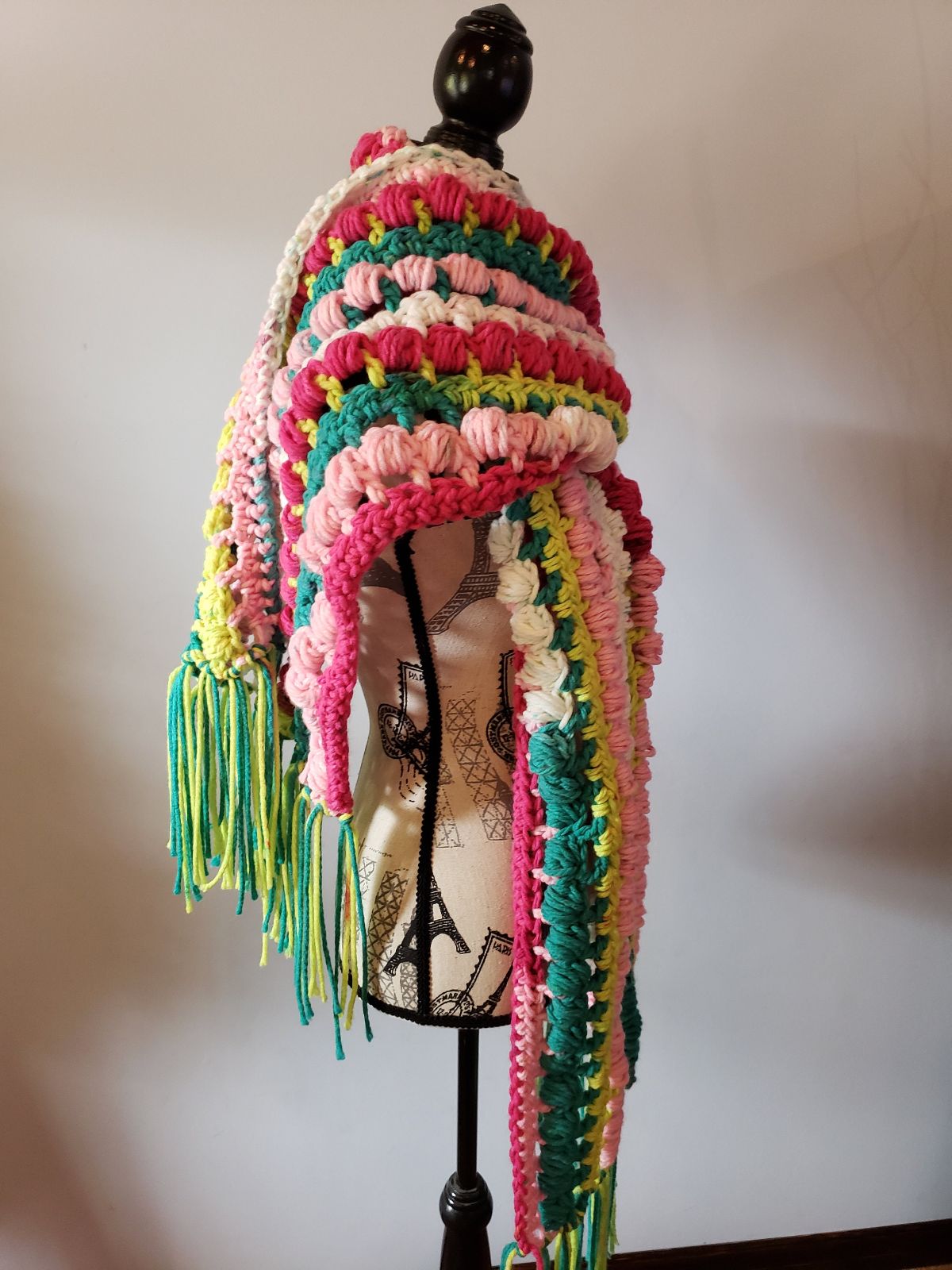 A mannequin wearing a chunky striped crochet scarf with pink, yellow, and green stripes and tassels on the bottom.
