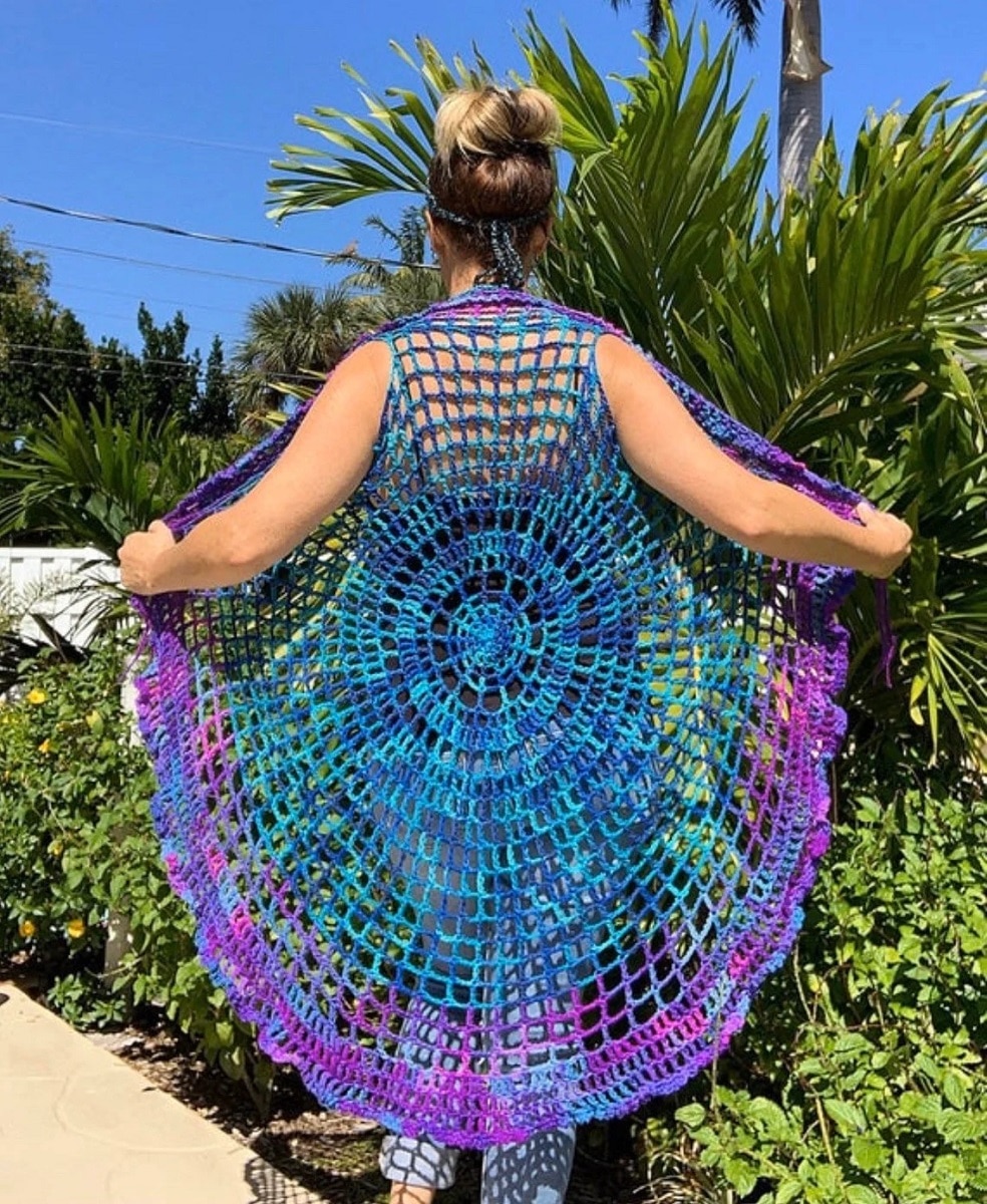 The back of a blond woman wearing a bright boho style crochet vest with purple, green, and blue circles of color across the loose stitched vest.