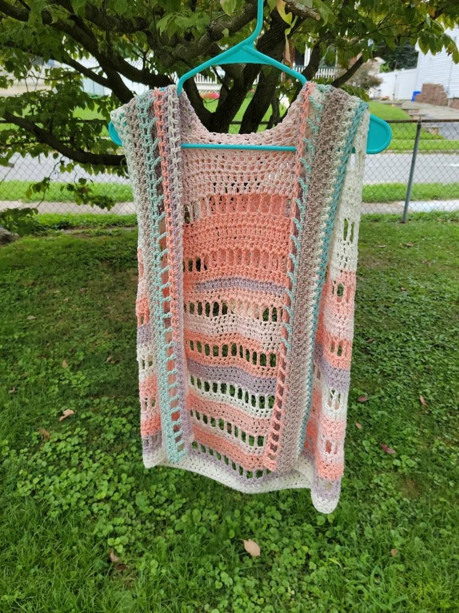  An open pink, green, orange, cream, and brown striped vest hanging on a green hanger from a tree above some grass.
