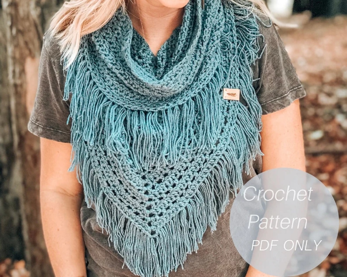 Teal crochet infinity scarf with gray and teal tassels hanging from all edges of the scarf worn by a blond woman.