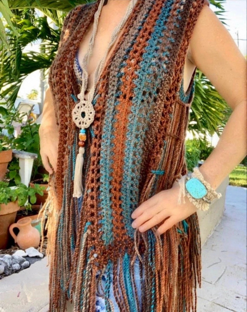 Woman wearing a long crochet vest with brown, orange, and blue vertical stripes and long tassels in these colors at the bottom of the vest.