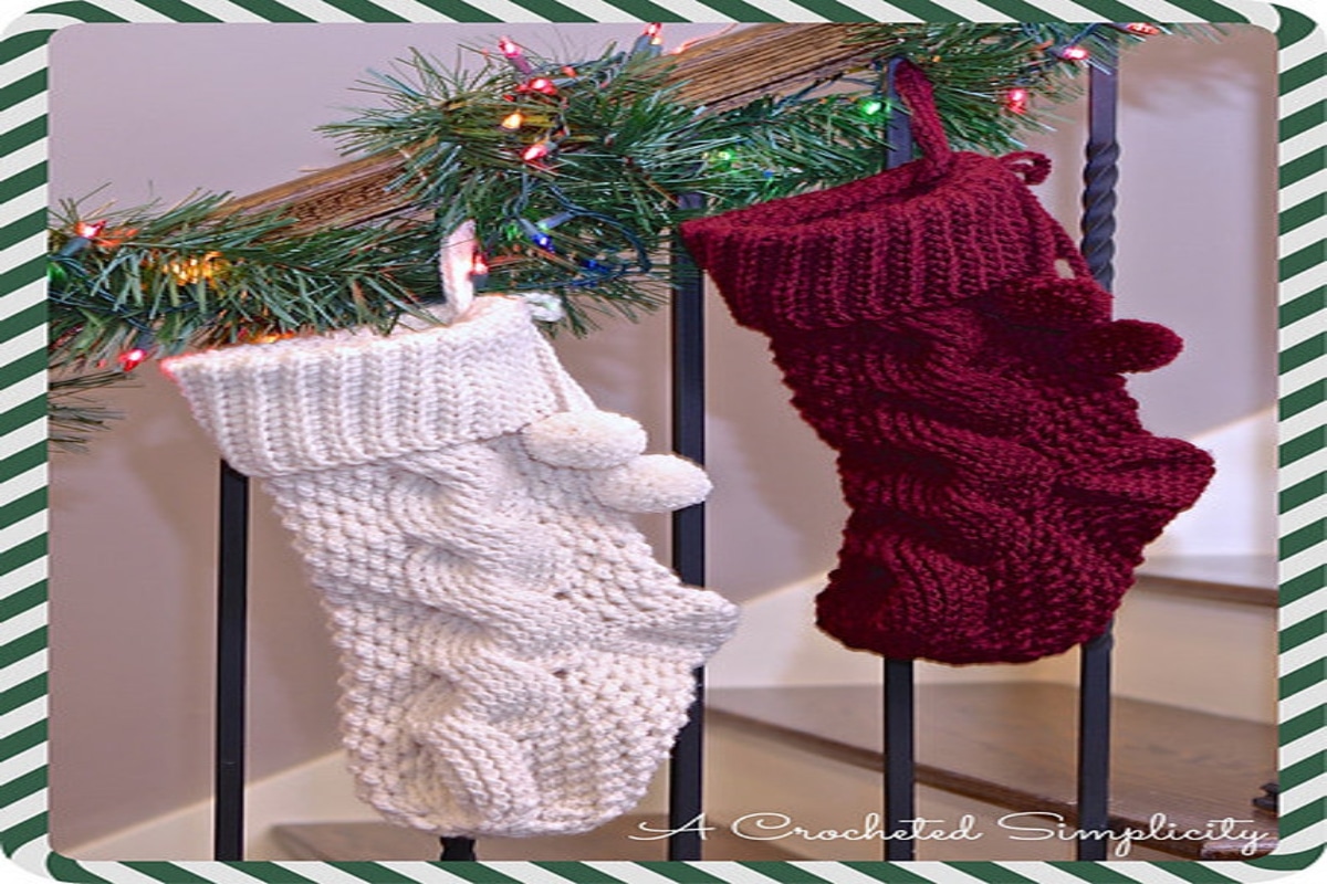 Red and white cable knit stockings with bobbles hanging off them attached to a banister decorated with pine and multi-colored lights. 