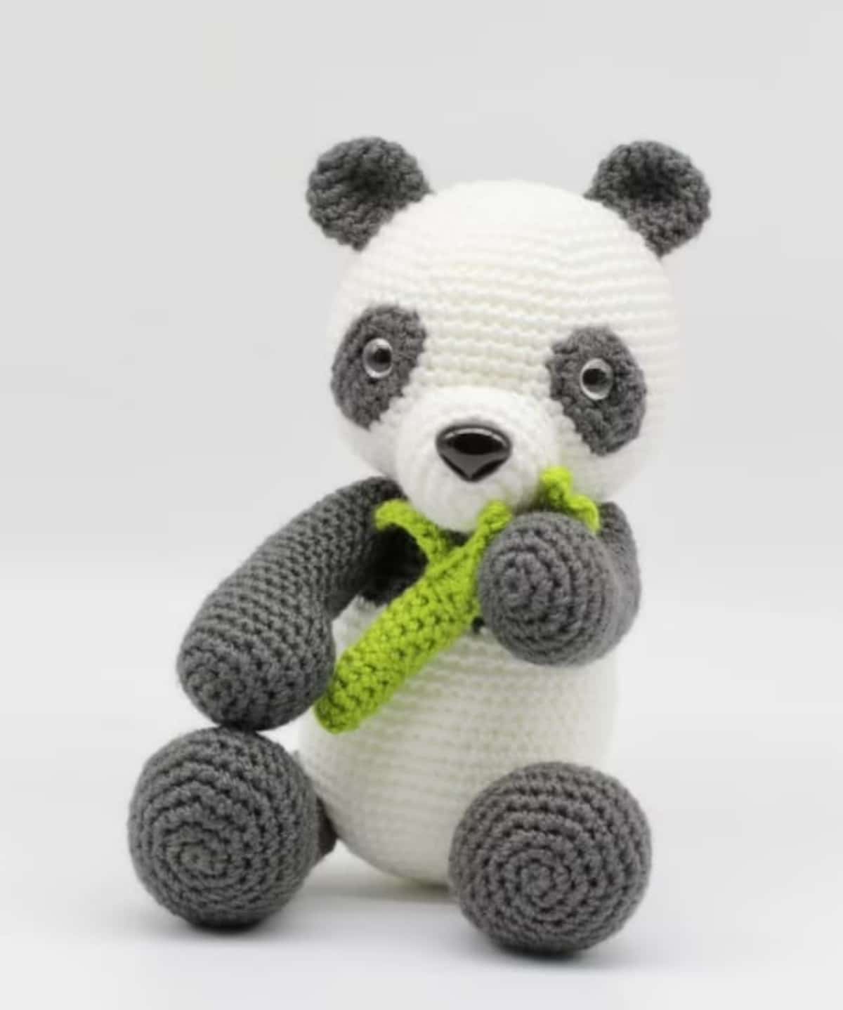 Black and white crochet panda bear sitting down with a green bamboo shoot in his arms and near his mouth on a white background.