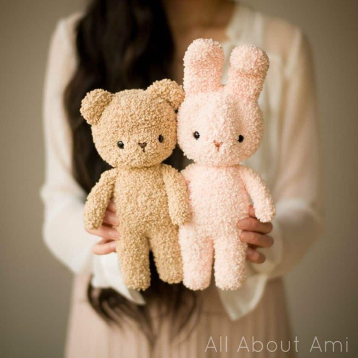 A dark haired woman holding a light brown crochet teddy bear and a pale pink crochet bunny on a white background.