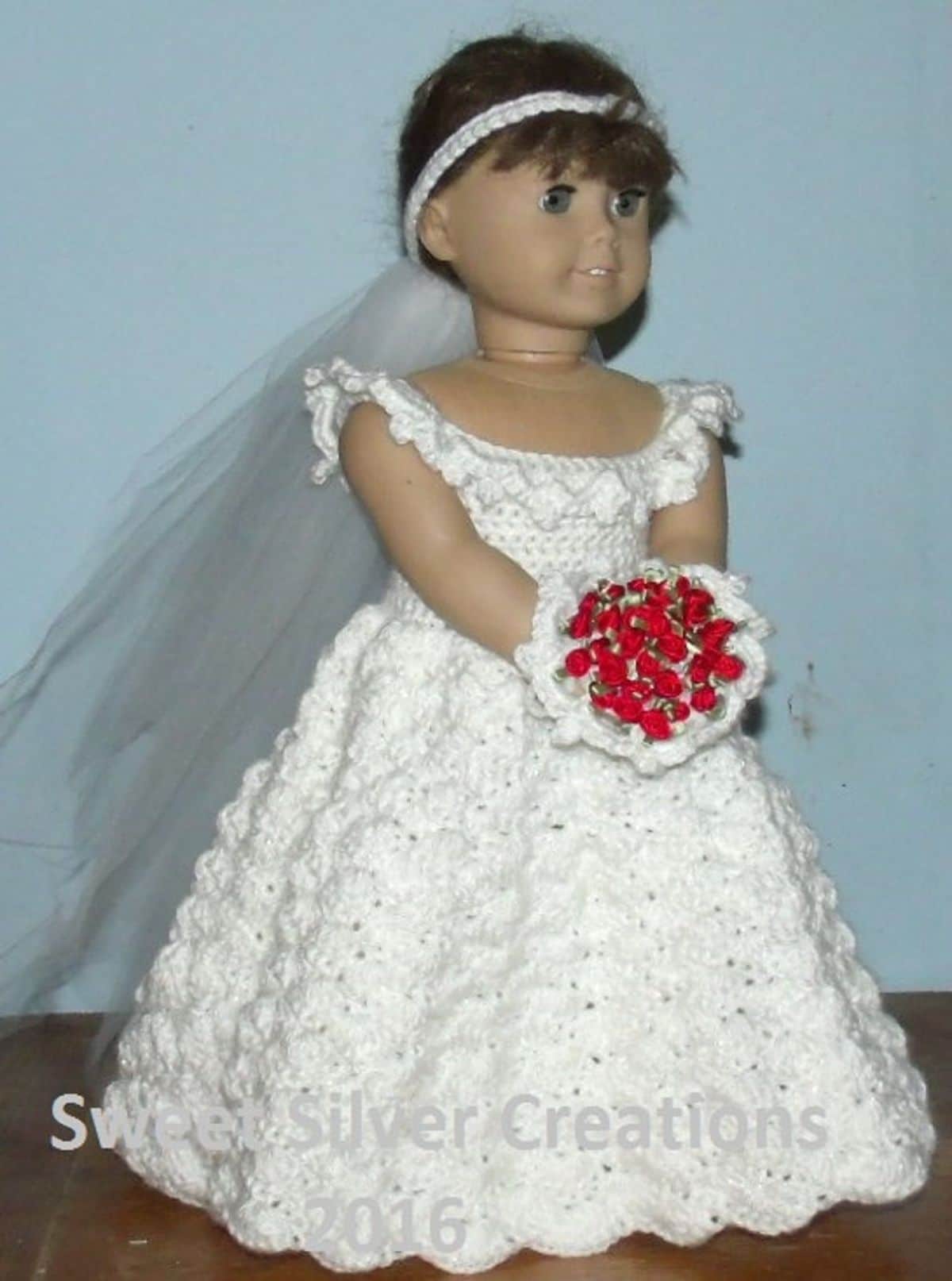 Brunette doll wearing a veil, crochet sleeveless wedding dress with a scoop neck and full skirt, holding a white and red bouquet. 