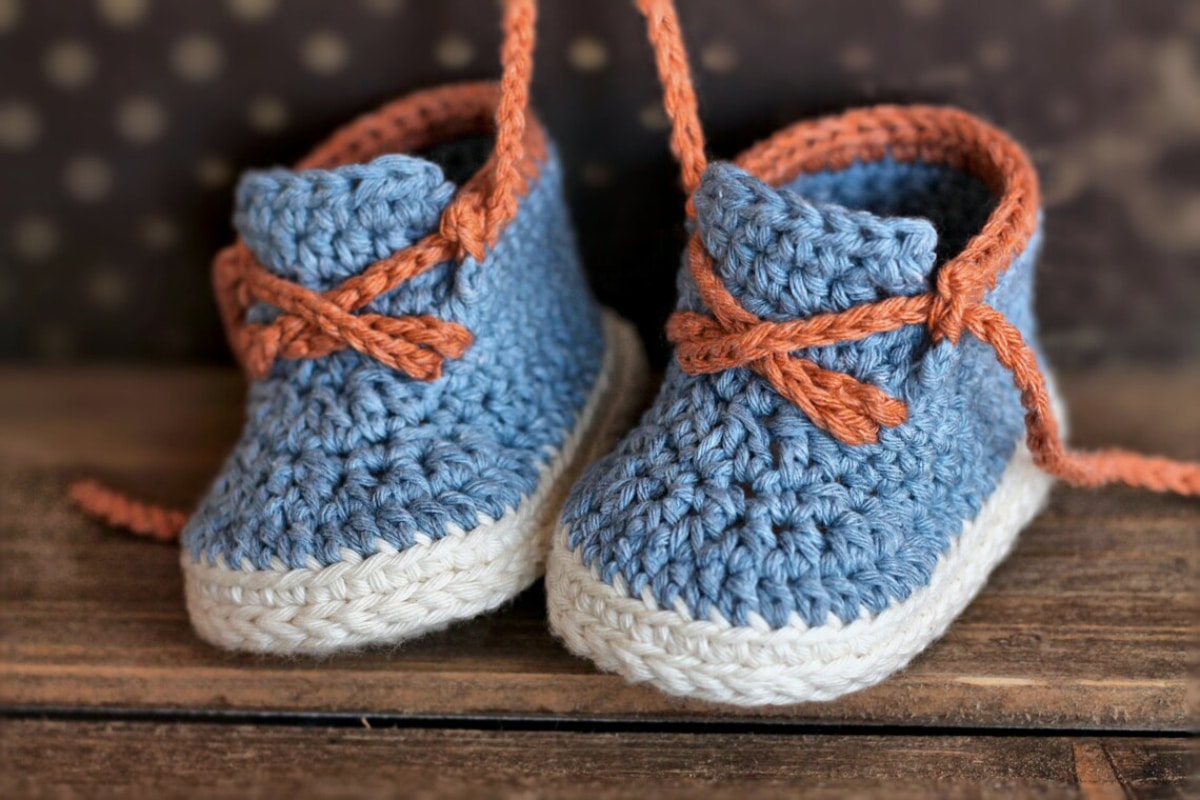 Blue crochet baby booties with white soles and brown laces to secure and a matching brown trim.