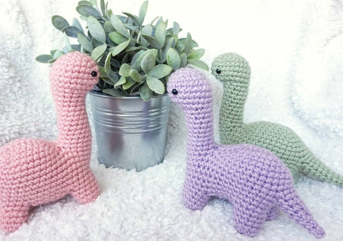 Pastel pink, purple, and green Brachiosaurus’ standing around a silver pot with an artificial plant on a white blanket.