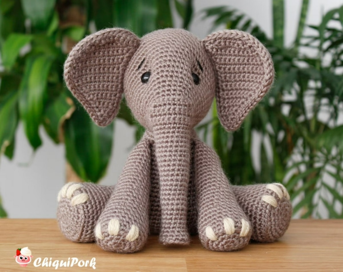 Large gray crochet elephant with white claws and small gray ears sitting on a wooden table in front of some plants.
