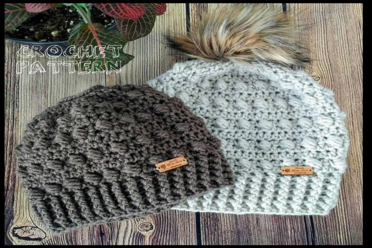 A small gray crochet messy bun hat next to a larger cream hat with a brown fluffy bobble on top on a wooden background.