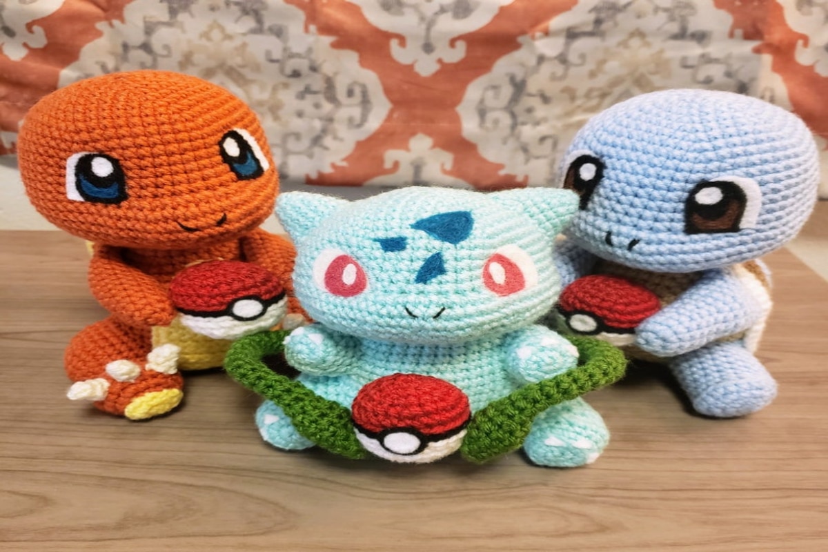 Stuffed crochet Bulbasaur, Charmander, and Squirtle sitting in a semi circle holding red, white, and black Pokeballs in their hands.