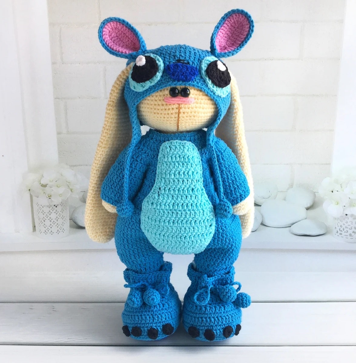 A cream waffle stitch crochet bunny wearing a blue Stitch costume standing on white wooden flooring.