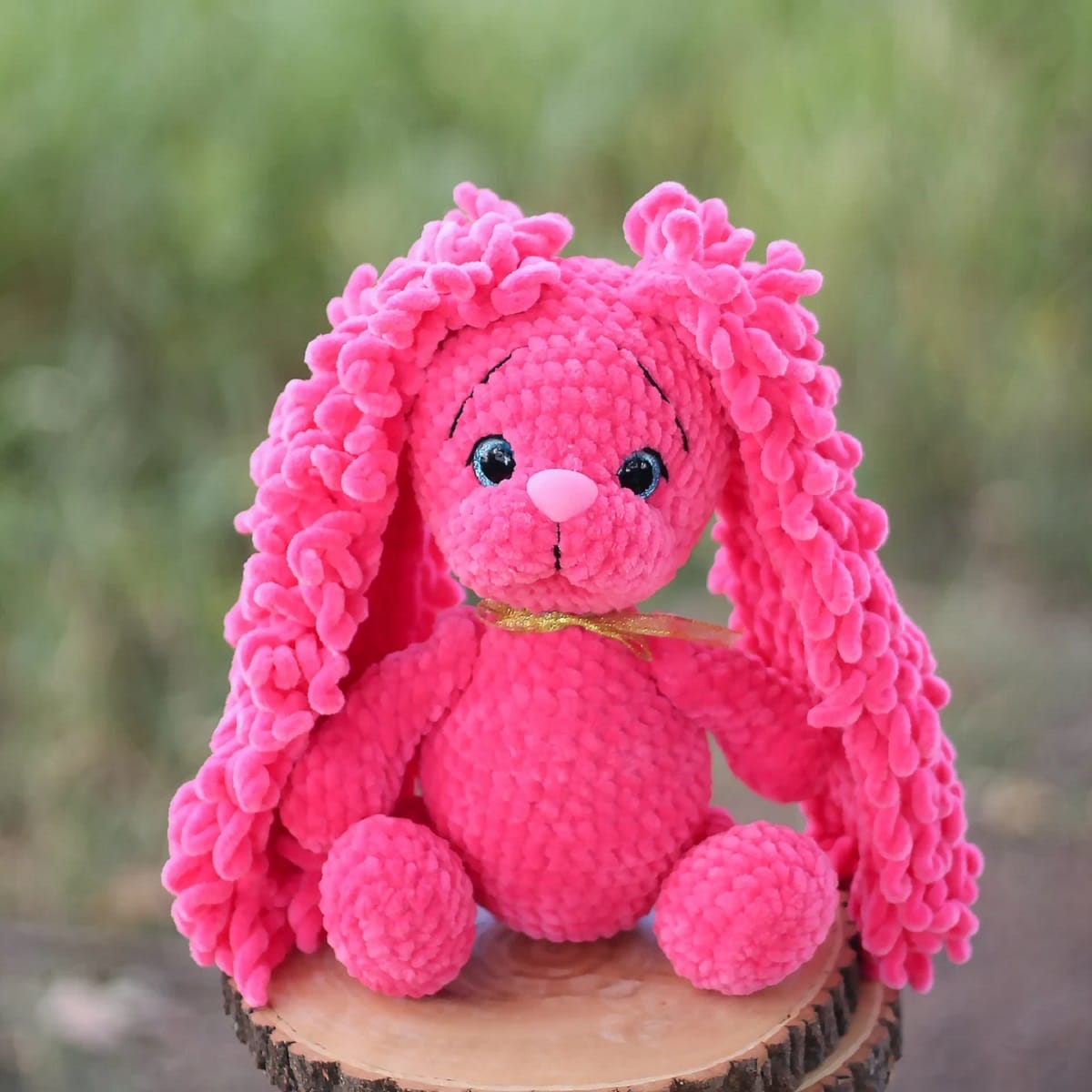 Pink crochet stuffed bunny you with oversized textured ears and a gold ribbon around its neck sat on a tree stump.