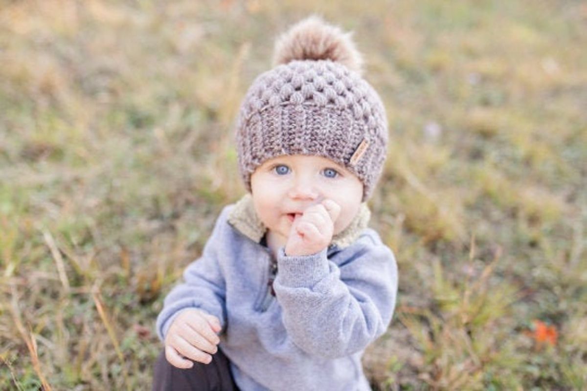 A baby sitting on grass wearing a gray crochet beanie with a brown fluffy bobble on top.