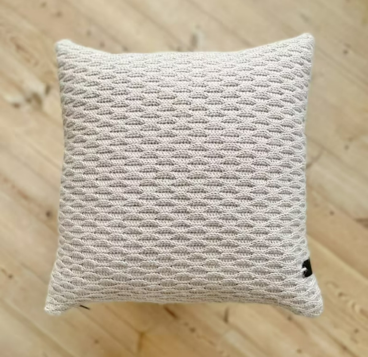 Cream crochet cushion with a textured 3-D effect on wooden flooring.