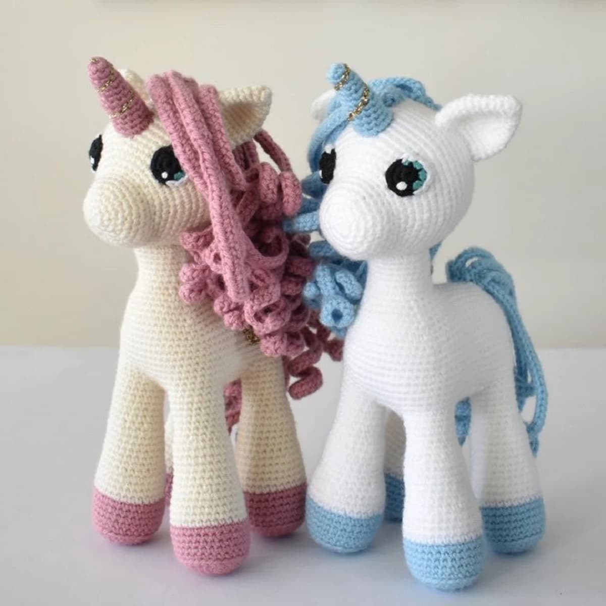 A cream crochet unicorn with a pink horn, mane, and feet next to a white unicorn with a blue horn, feet, and mane.