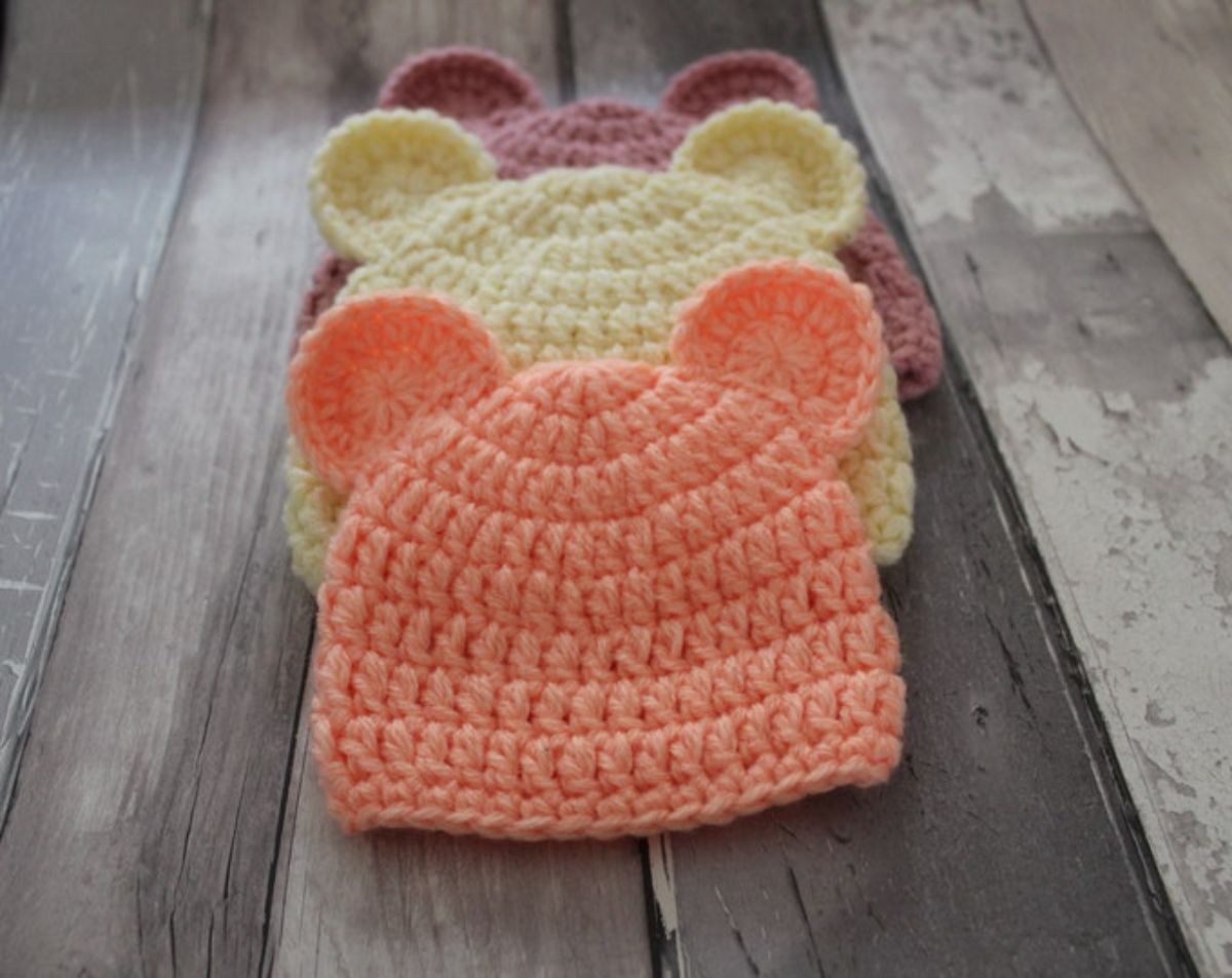 Salmon, yellow, and purple beanies lying on a wooden floor with small teddy bear ears stitched on the side of each beanie.