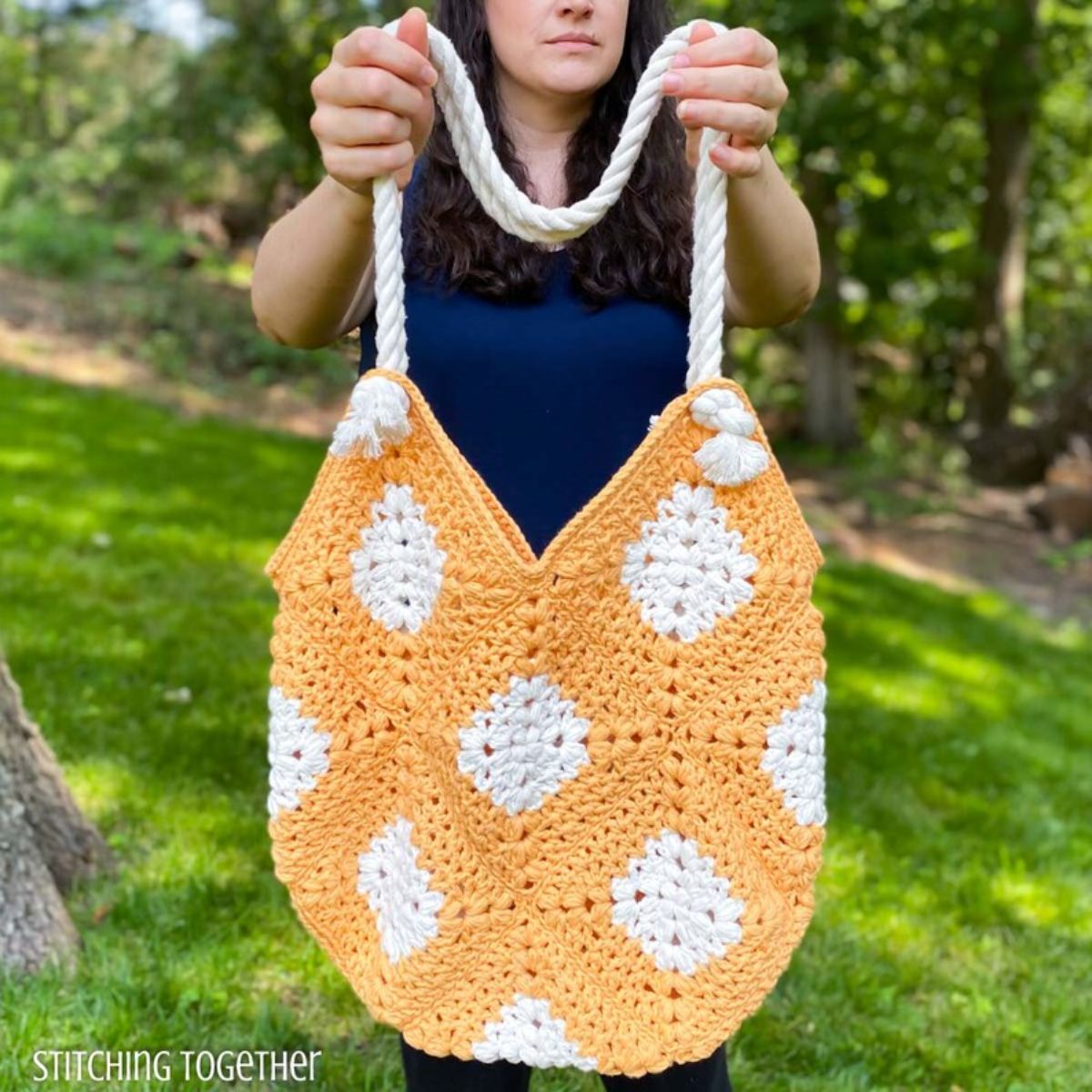 Brunette woman holding a yellow and white diamond style crochet market bag with two thick, rope-like handles. 