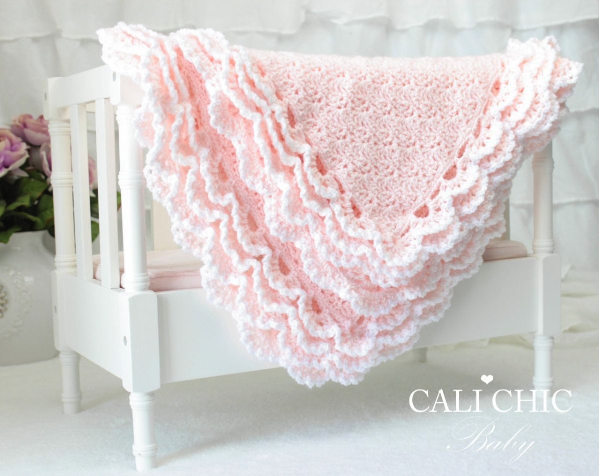 Small baby pink crochet baby blanket with a thick white lace trim folded over the railings of a white crib.