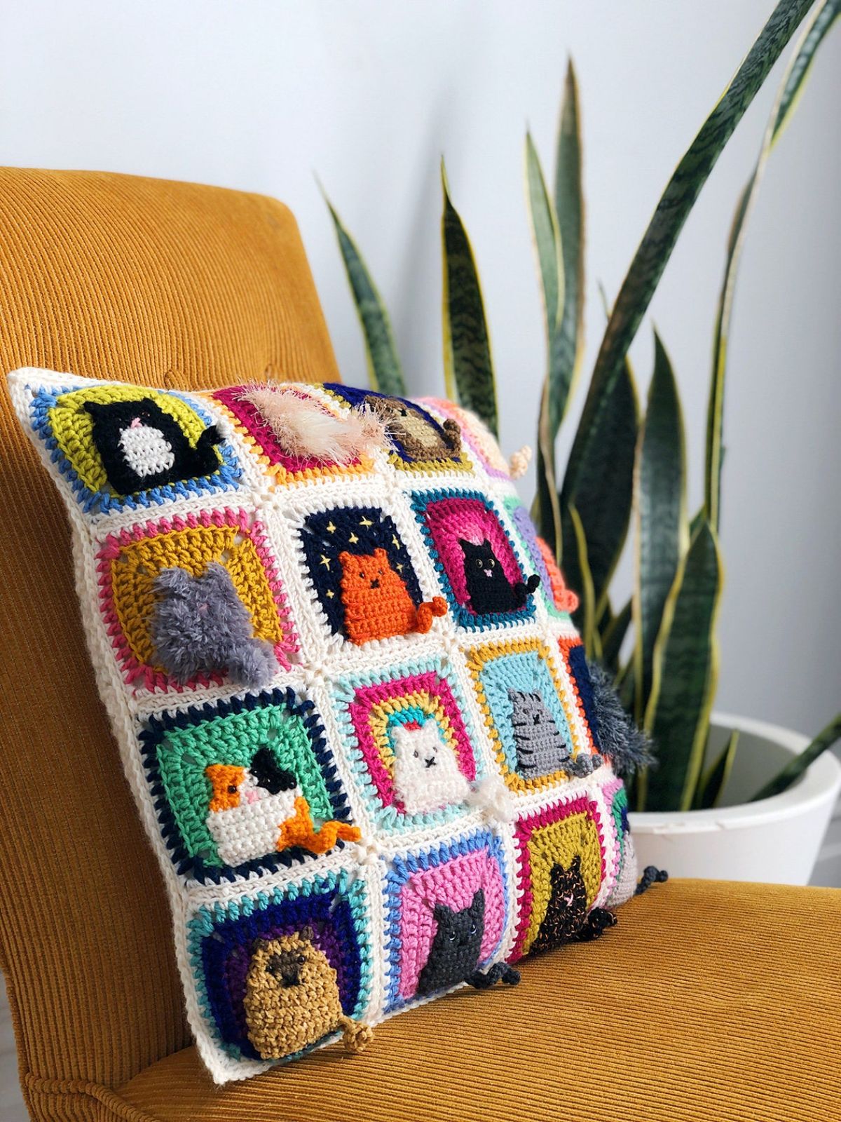 White crochet cushion with sixteen bright colored squares with a different cat stitched into each square on a yellow chair.