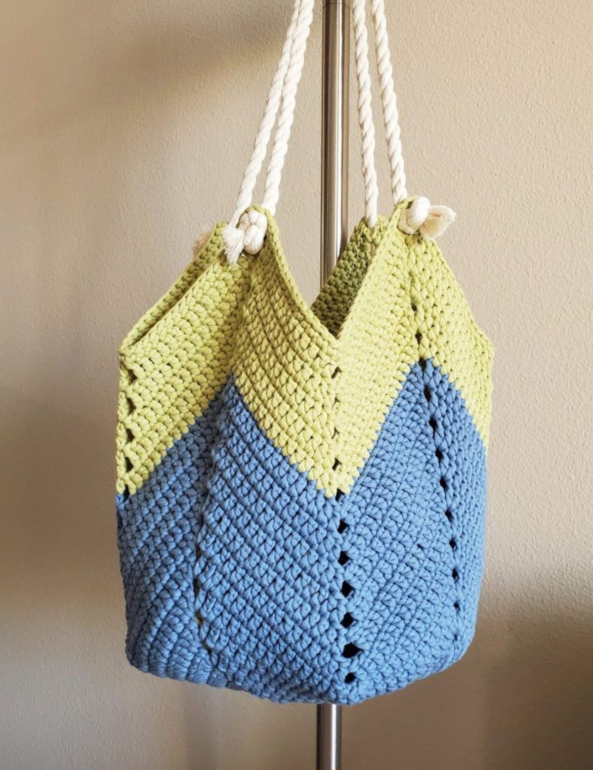 Large blue and yellow crochet chevron shoulder bag with thick white rope for the straps hanging from a metal pole.