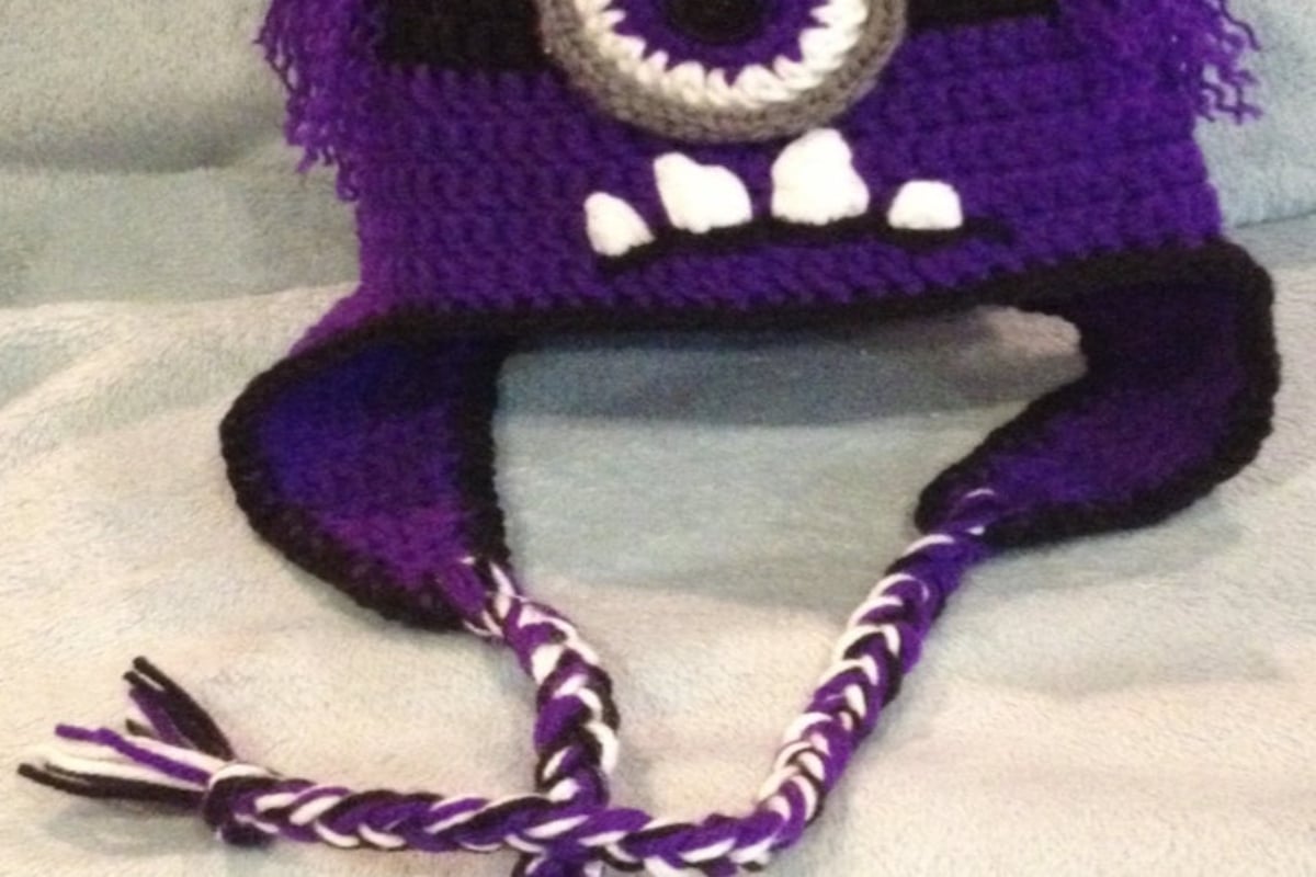 Dark purple evil minion hat with white and black eye, wonky teeth and purple and white braids on the ear flaps.