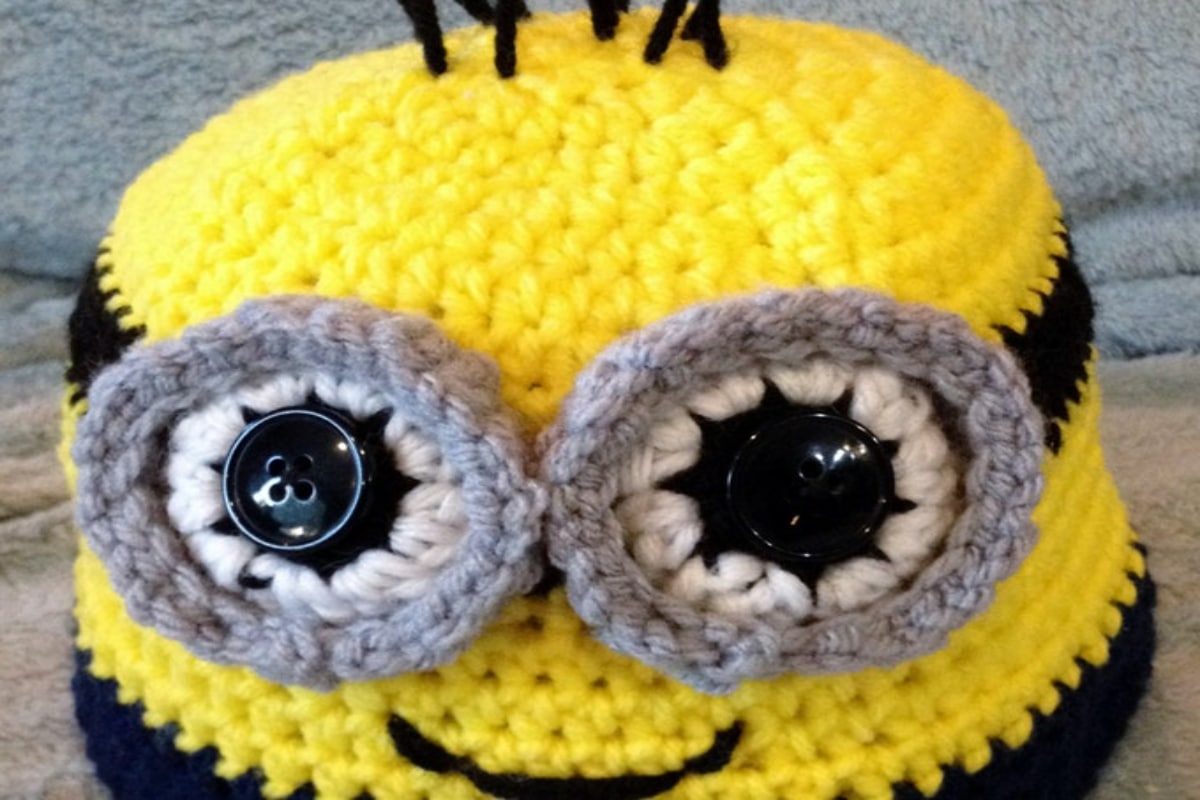 Yellow crochet minion style hat with black, white, and gray eyes, a smiling mouth, and black spikes on top as hair.