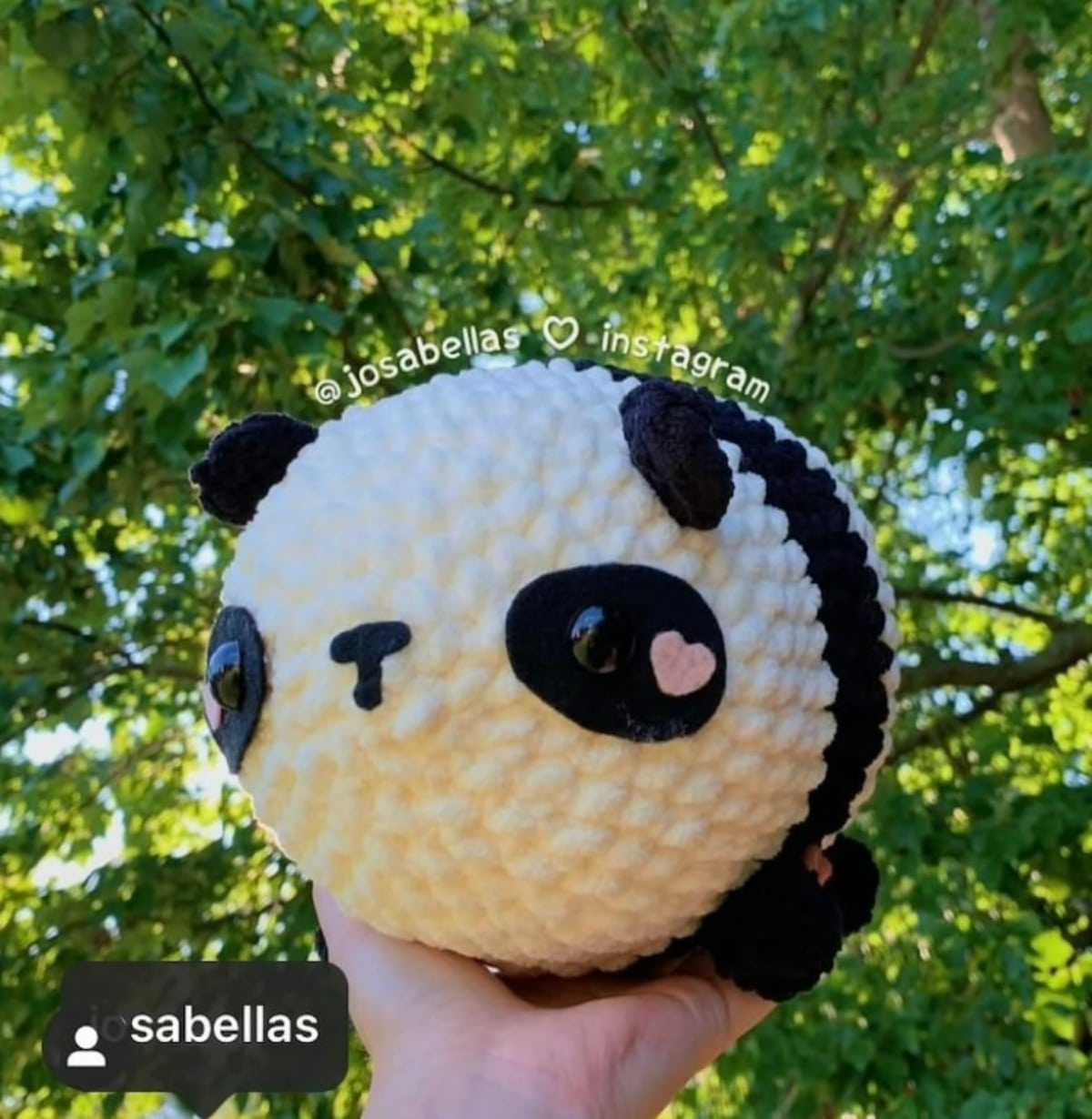 Large oval shaped crochet panda with pink hearts next to its eyes and a bobbled effect on its body held next to a tree. 