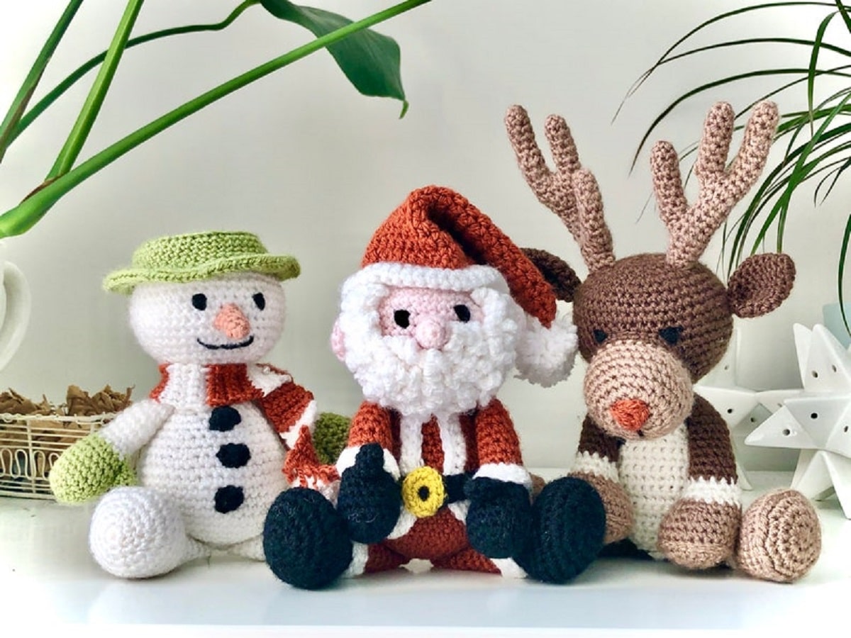 A crochet snowman sitting next to a Santa and reindeer on a white background with green house plants behind them.