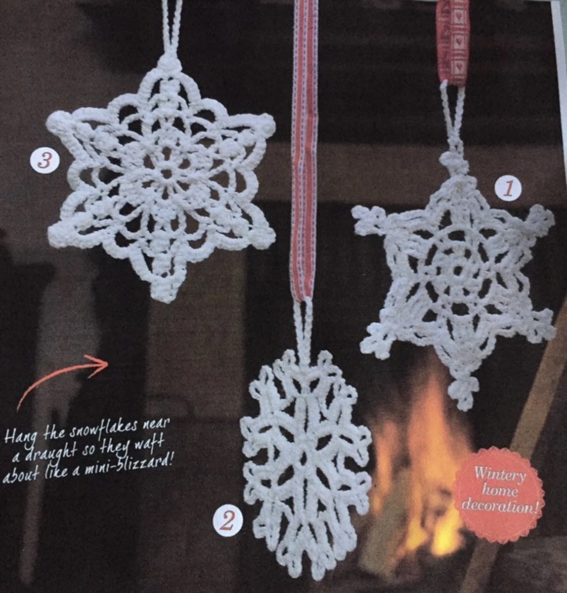  Three white crochet snowflakes hanging from white and red ribbon in front of a roaring fire.