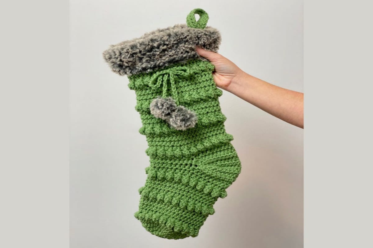 Green Christmas stocking with small balls in rows and gray fluffy banding. Two matching gray bobbles sit in the center.