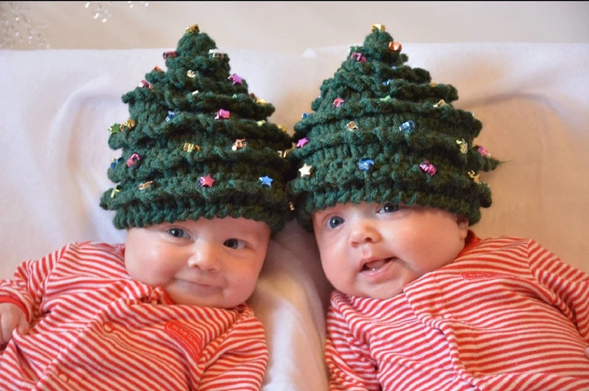 Two babies lying next to each other in green crochet Christmas hats with small circles stitched in to look like lights.