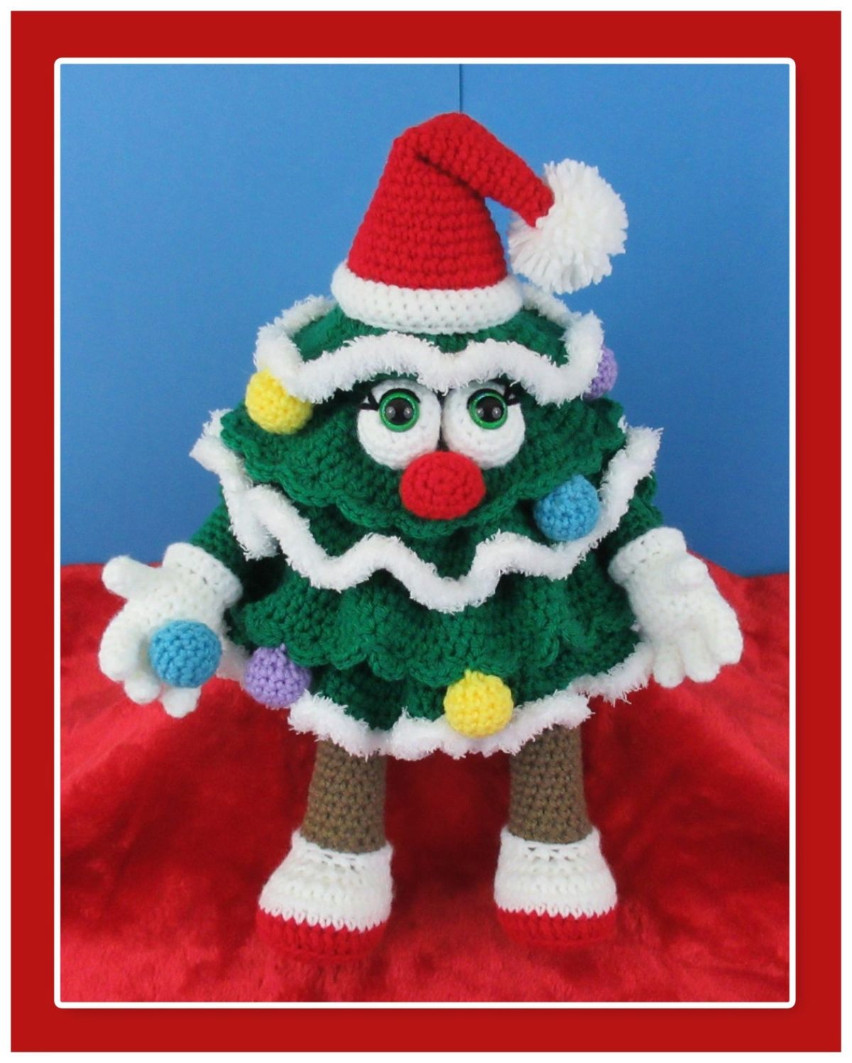A large green crochet Christmas tree with white edges, white hands, and brown legs wearing a Santa hat and red shoes.