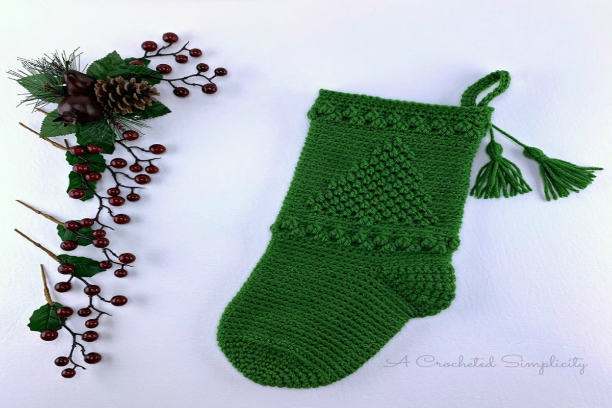 Green crochet stocking with Christmas tree design knitted into it. Stocking has two tassels and is next to some holly.