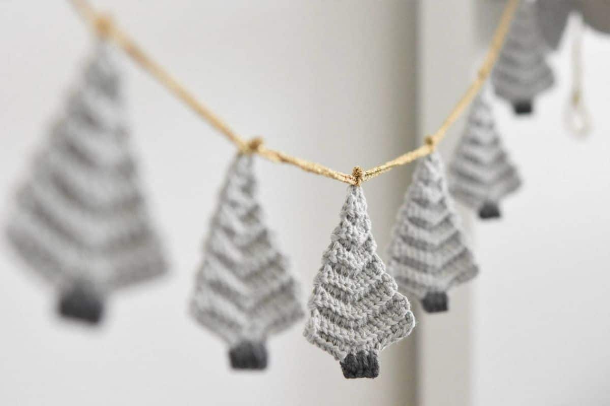 Gray crochet Christmas tree bunting hanging on a brown rope by a white wall.